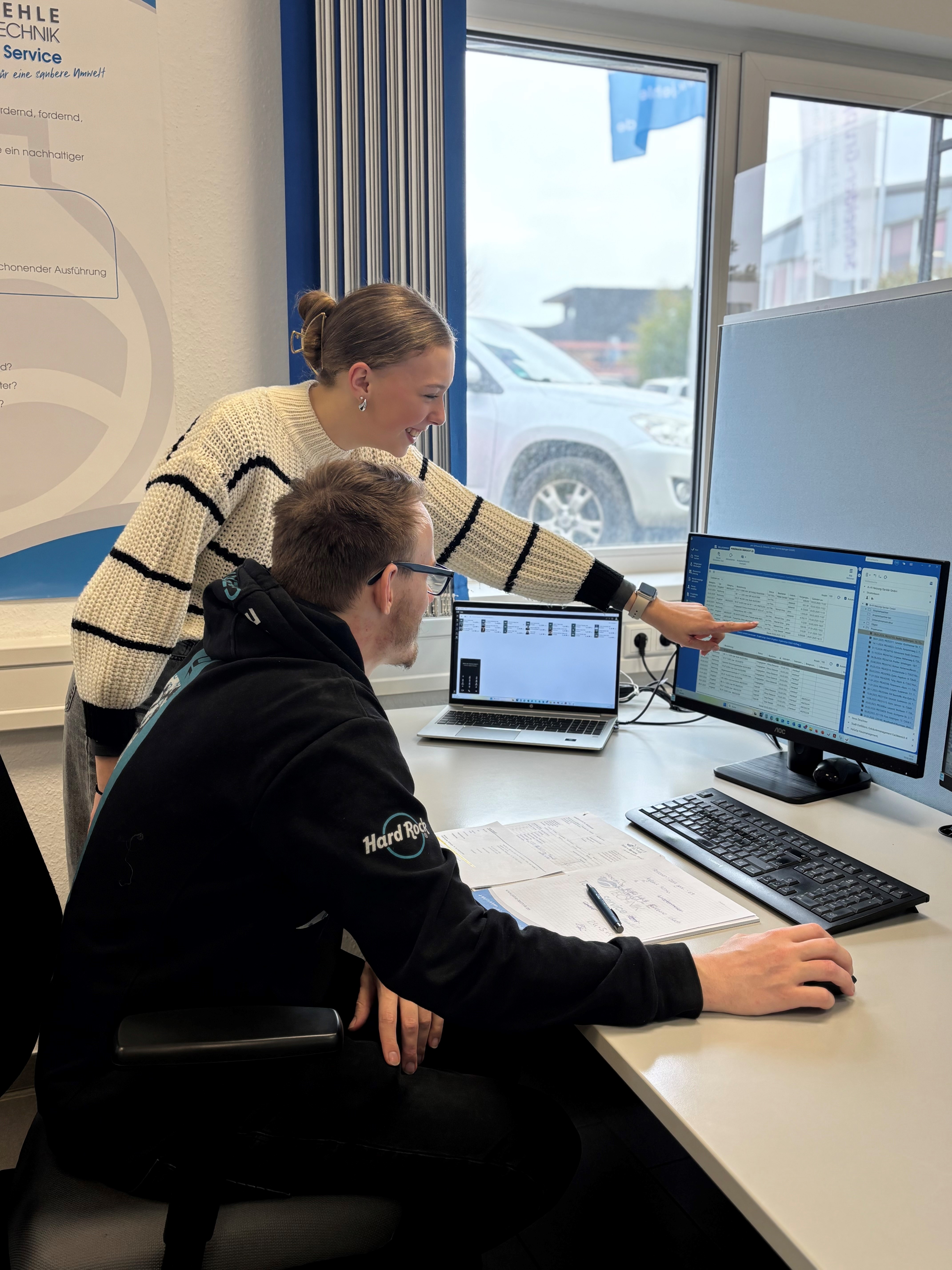Frau zeigt auf Monitor, Mann sitzt vor Bildschirmen, wirkt konzentriert.
