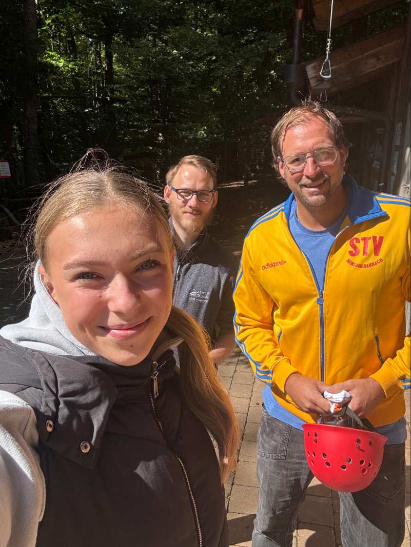 Eine junge Frau lächelt selbstbewusst, zwei Männer stehen dahinter in gelber und grauer Jacke