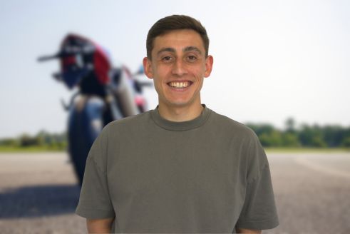 Mann mit neutralem Blick in grauem Shirt vor rotem Motorrad im Unscharfen